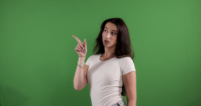 Young woman pointing with finger on green screen background, looking surprised
