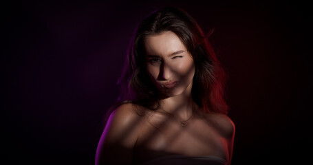 Naklejka premium Studio portrait of a young woman with long brown hair, winking and pouting, illuminated by purple and red lights casting shadows on her