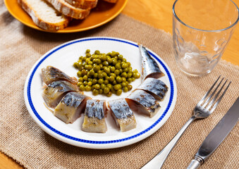 On checkered tablecloth there is plate with dinner of sea fish - fried mackerel scomber carcass fillet, complemented with edible podded pea green peas