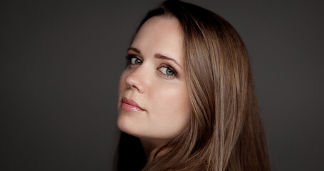 Studio portrait of a young woman with long brown hair looking over her shoulder against a gray background
