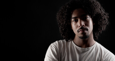 Portrait of a serious young African American man with curly hair posing in a studio against a black...