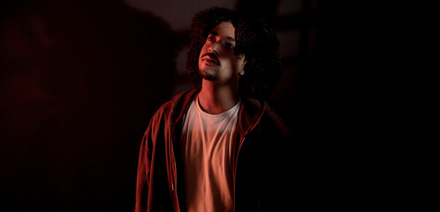 Portrait of a stylish young man with curly hair looking up, illuminated by a red light in a dark...
