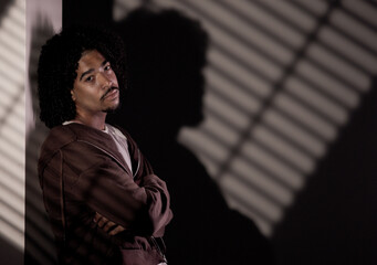 Pensive young adult male leaning against a wall with arms crossed and striped shadow