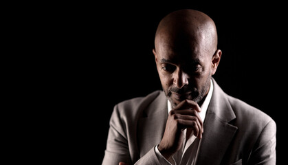 Low key portrait of thoughtful businessman with hand on chin, looking down against black background