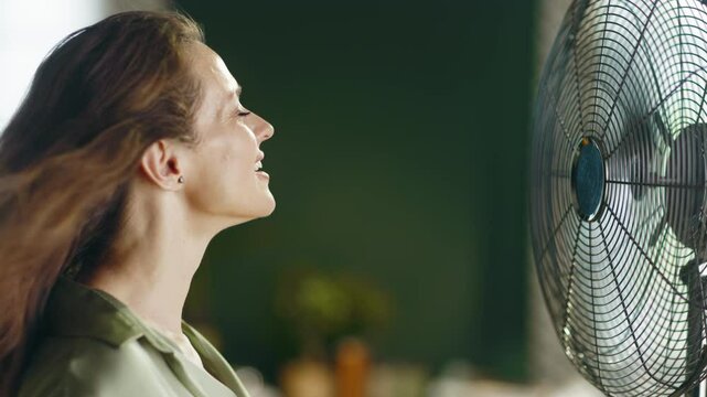 A smiling young woman finds refreshing relief from summer heat as an electric fan gently blows her long brown hair, creating a sense of comfort and relaxation against a dark green background.