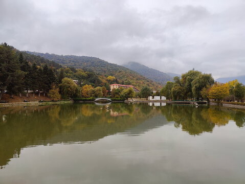 A tranquil lake reflecting colorful autumn trees and a forested mountain on a cloudy day. Scenic park landscape with a small bridge.