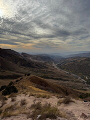 Dramatic mountain landscape view from a high vantage point. Arid hills and a valley with a road under a cloudy sky.