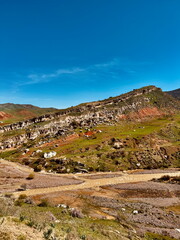 Rugged natural landscape featuring a muddy braided river. Layered geological formations on a green hillside under a vast blue sky.