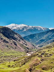 A stunning mountain landscape featuring a lush green valley, a winding river, and snow-capped peaks under a clear blue sky.