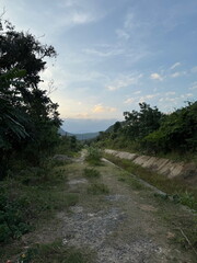 A rural dirt path winds through a lush green valley towards distant mountains at dusk. Scenic countryside trail next to an irrigation canal.