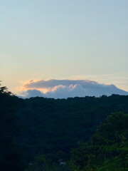 Naklejka premium Peaceful mountain landscape at dusk. Glowing clouds from the sunset surround a distant peak above a lush green forest.