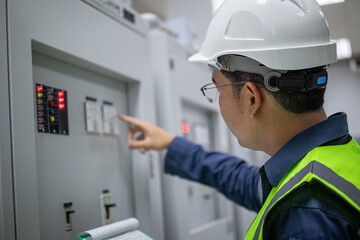 Electrician wearing safety helmet and vest performs control panel inspection while engineer points...