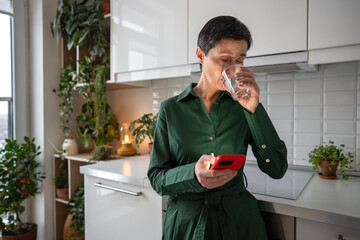 Elderly woman drinking water following smartphone reminders and schedule. Hydration maintenance,...