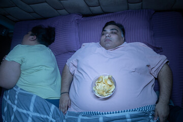Overweight indian couple with unhealthy junk food while sleeping in bed. Plus size husband wife resting together.