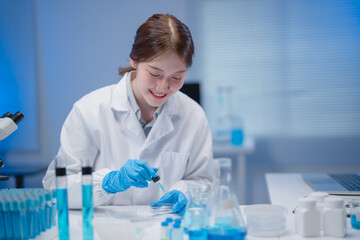 Young asian female scientist wearing lab coat and gloves dropping blue liquid in petri dish using pipette, doing research in a modern laboratory with microscope and test tubes