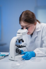 Scientist wearing gloves and a lab coat closely examines samples through a microscope in a...