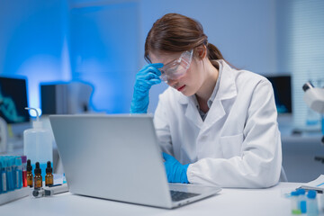 Young scientist wearing lab coat and gloves is feeling tired while working with her laptop in a...