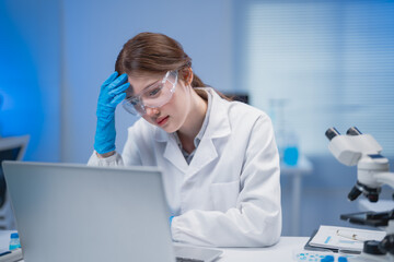 Young female scientist wearing lab coat and protective glasses feeling tired and stressed while...