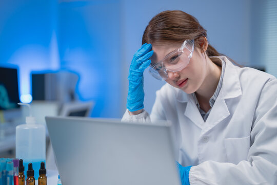 Young scientist suffering from burnout syndrome while working on laptop in modern laboratory with blue light, she's touching her head with gloved hand