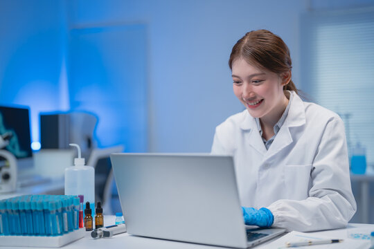 Young female scientist wearing lab coat and gloves is working with laptop in modern laboratory, analyzing medical research data and smiling, with test tubes and microscope in background - Powered by Adobe