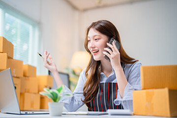Smiling young woman managing her online business, talking on the phone while working on a laptop. Surrounded by packages, she embodies the modern e-commerce entrepreneur