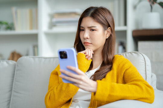 serious young asian woman sitting on sofa and looking at mobile phone screen feels Upset,sad and disappointed.Unhappy asian female receive bad news,black message while sitting on couch at home