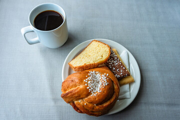 Syrup, glazed, huge cinnamon, cardamom, brown, braided bun (Kanelbullar), accompanied by chocolate and shortbread biscuit cookies and pound cake topped by white pearl-sugar sprinkles with coffee cup