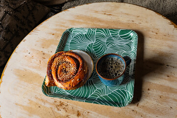 Delicious, sweet, glazed, large cinnamon and cardamom spiced spiral pastry bun (Kanelbullar or Kanelbulle), topped with syrup and white pearl-sugar sprinkles, with hot cup of coffee in a rustic scene