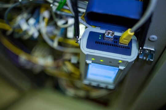 Handheld tester displaying diagnostics with green LEDs, yellow cable, patch cables inside rack