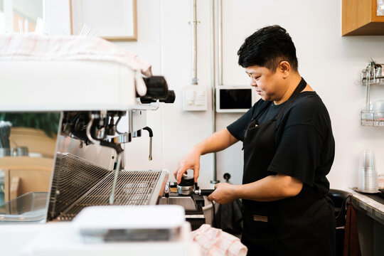 Focused barista prepares fresh coffee behind espresso machine in a cozy cafe. Daily work routine highlights dedication and quality service in small business coffee shop environment.