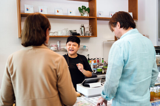 Cafe owner explains menu to customers at counter in modern coffee shop. Small business engagement, friendly service, and hospitality in a welcoming local setting with diverse interaction.