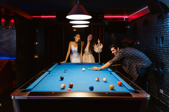 Three stylish friends enjoy a game of pool in a trendy lounge. Laughter fills the air as one lines up a shot, creating a fun and social moment in a vibrant nightlife setting.