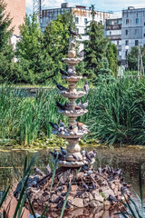 Multi-tiered stone fountain covered with pigeons in Heroes of Ukraine Square, Lokhvytsia, Poltava region, Ukraine. Urban park water feature surrounded by green vegetation and residential buildings