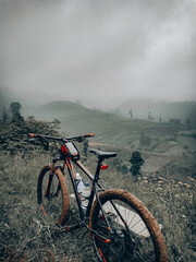 Obraz premium A scenic view from a bridge shows a mountain bike resting against a red and black railing. In the background beautiful rice field views under a foggy weather.