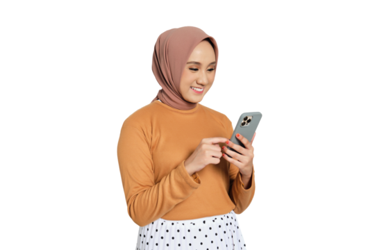 Cheerful young Muslim woman in hijab texting on smartphone while standing isolated on transparent background