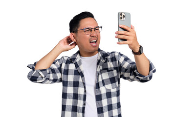 Confused young man wearing glasses scratching head and looking at smartphone screen with puzzled...
