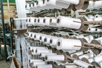 Industrial Weaving in Action – Large-Scale Yarn Processing and Fabric Roll Production Inside a Textile Factory. Industrial Textile Weaving Machines in a Factory. Spools of white thread in a textile.