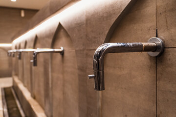 A row of minimalist chrome ablution taps stand out from the brown textured wall, illuminated by...