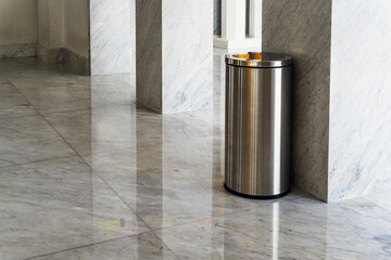 A clean cylindrical stainless steel trash can stands in the corner of the shiny marble floor,...