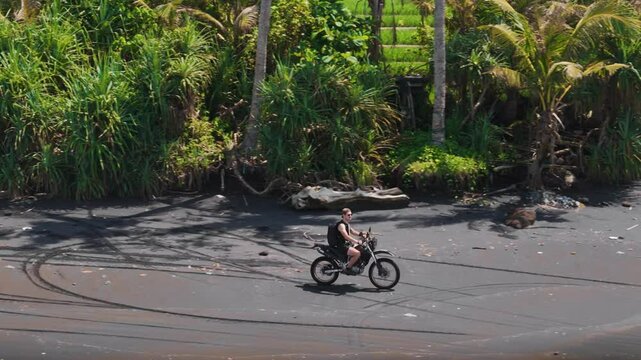 Fast parallel drone tracking shot alongside a dirt bike rider speeding along the black sand beach at Pasut, Bali. Tropical vegetation and tire marks add texture to this thrilling adventure moment