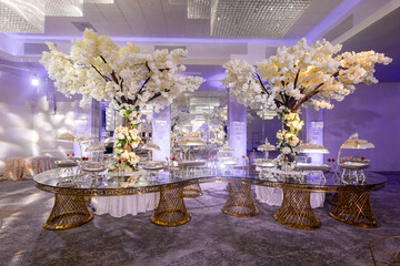 Luxurious wedding reception interior with blooming white flowers trees and crystal chandeliers....
