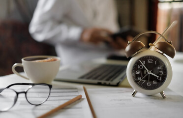 select focus on alarm clock on table near laptop, documents and eyeglasses and blur businessman working behind, time management concept, routine work, office hour