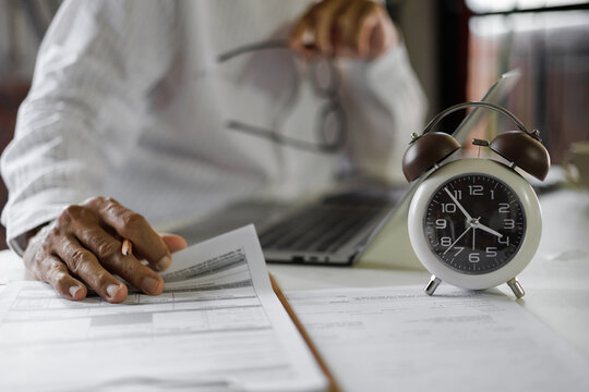 selective focus on alarm clock, asian businessman review business document using laptop working on table before work time end in a day, time management concept, routine work, office hour - Powered by Adobe