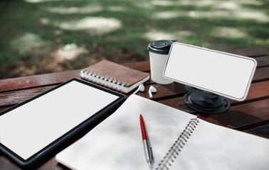 mobile phone on stand with coffee cup, digital tablet and notebook on wooden table in park, working outside concept and relax via modern technology from mobile