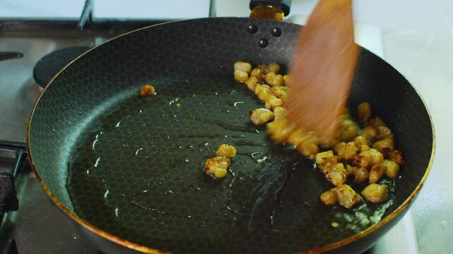 Stirring finely chopped fat pieces in a frying pan with a wooden spatula. Close-up of fat frying and rendering in a hot skillet in home kitchen