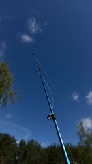 Close-up of blue fishing rod against clear summer sky — outdoor angling gear with visible fishing...