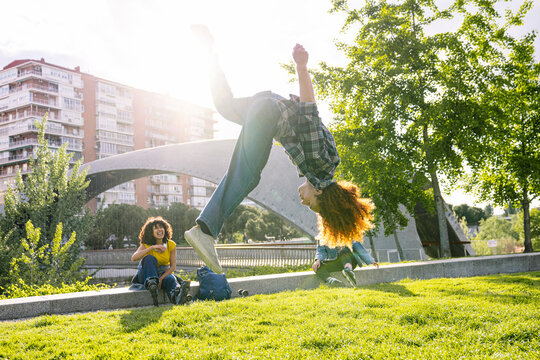 Redhead skater performing backflip in urban park while two friends watch, enjoying leisure time on sunny day - Powered by Adobe
