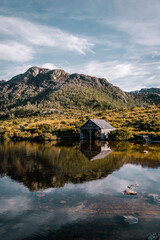 A rustic wooden hut sits by the edge of Love Lake with Cradle Mountain in the background, its...