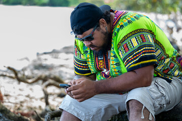Latin american tourist using mobile phone sitting on a trunk on the beach in Cahuita National Park,...