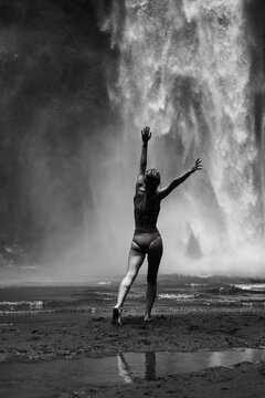 Woman in swimsuit reaches skyward beside thundering tropical waterfall